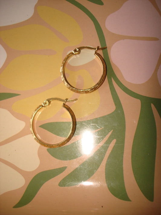 A pair of gold hoop earrings with a textured, hammered finish, displayed on a retro floral-patterned surface.